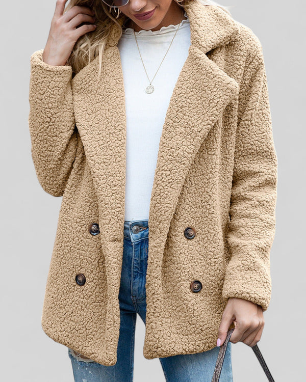 Vrouw draagt een trendy, beige teddyjas met dubbele knopen, gecombineerd met een witte trui en spijkerbroek. Perfect voor herfstmode en casual stijl.