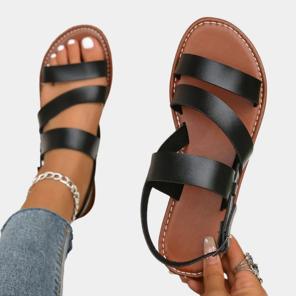 Zwarte leren sandalen met bruine zool, gedragen door een persoon met jeans en enkelbandje. Stijlvolle zomerschoenen voor dames.