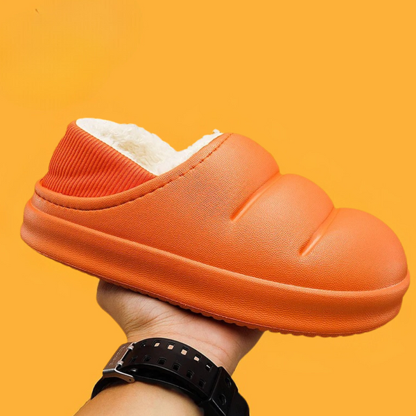 Oranje pluche pantoffel met dikke zool, vastgehouden door een hand, tegen een oranje achtergrond. Comfortabele huispantoffel voor dagelijks gebruik.