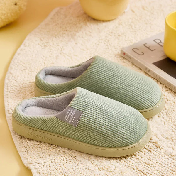 Groene ribfluwelen pantoffels op beige tapijt, naast een geel kopje en tijdschrift. Comfortabele, warme huisschoenen voor dagelijks gebruik.