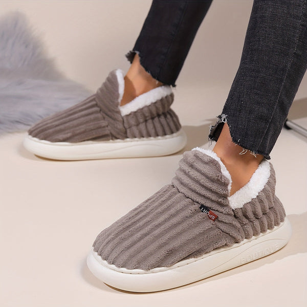 Grijze pluche pantoffels met witte voering, gedragen door persoon in zwarte jeans. Comfortabele winterse huisschoenen voor dames.