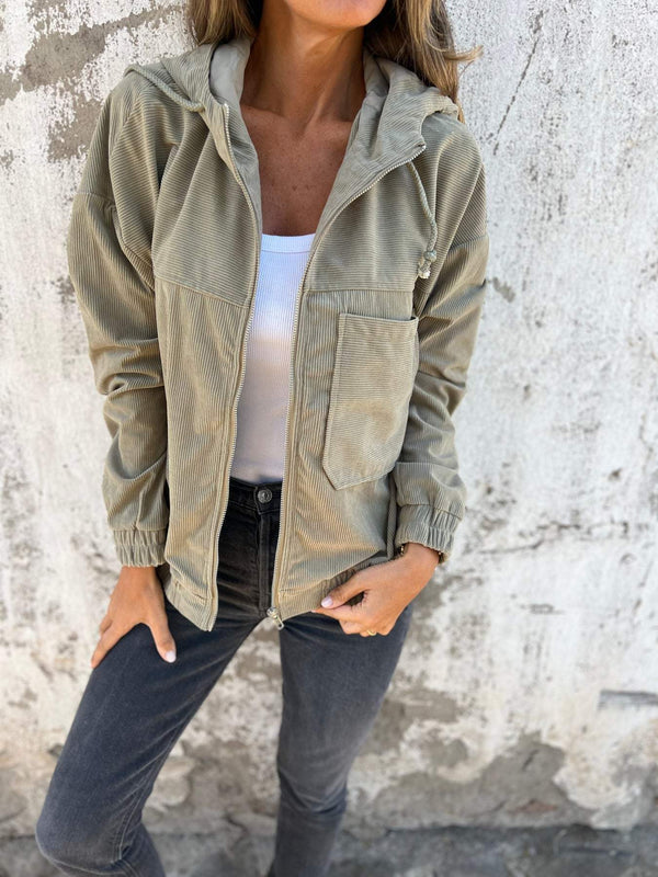 Vrouw in beige corduroy jack met capuchon, witte top en donkere jeans tegen verweerde muur. Modieuze herfstkleding, casual damesstijl.