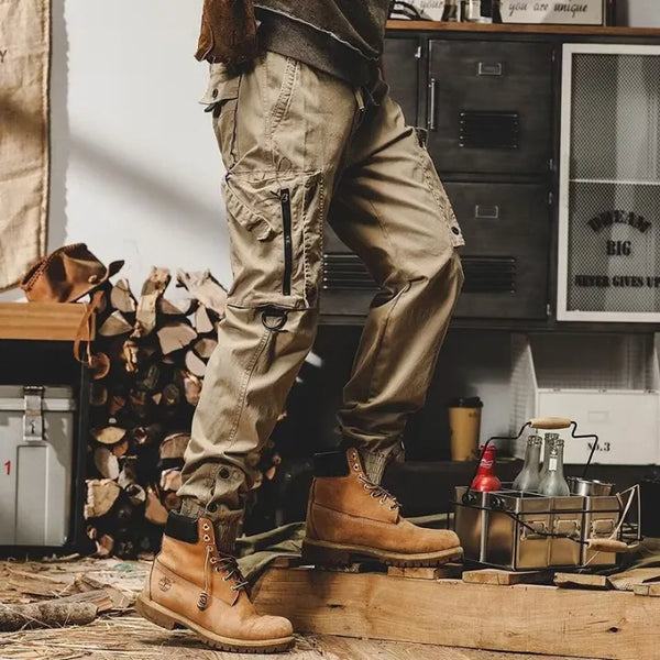 Man in beige cargobroek en bruine werkschoenen in een rustieke werkplaats met houtblokken en metalen kisten. Outdoor kleding, robuuste stijl.