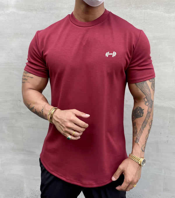 Man in bordeauxrood sportshirt met halterlogo, getatoeëerde armen, gouden sieraden, en mondkapje. Fitnessmode, casual herenkleding.