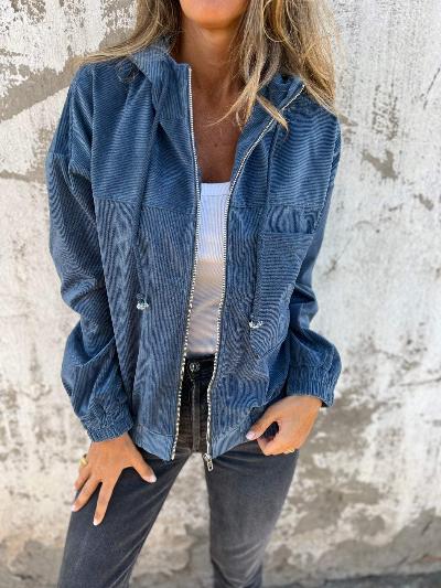 Vrouw in blauwe corduroy jas met rits, witte top en zwarte jeans tegen betonnen muur. Modieuze dameskleding, casual herfstoutfit.