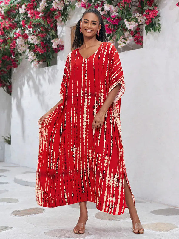 Vrouw in rode kaftan jurk met tie-dye patroon, staand voor witte muur met bloemen. Zomermode, bohemian stijl, comfortabele dameskleding.