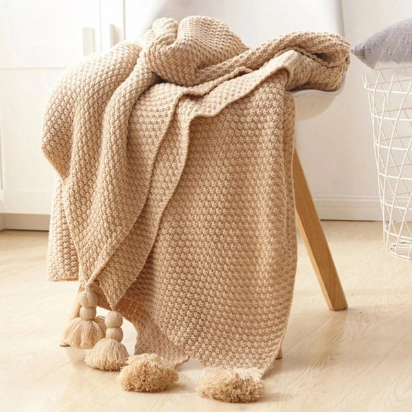Beige gebreide deken met pompons, gedrapeerd over een witte stoel. Perfect voor interieurdecoratie, woonkamer, slaapkamer, gezellig en stijlvol.