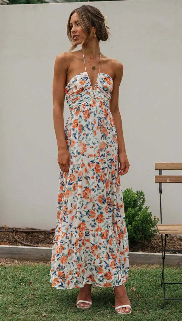 Vrouw in lange, mouwloze zomerjurk met bloemenprint, halternek en sandalen, staand in tuin. Perfect voor zomermode en casual buitenkleding.
