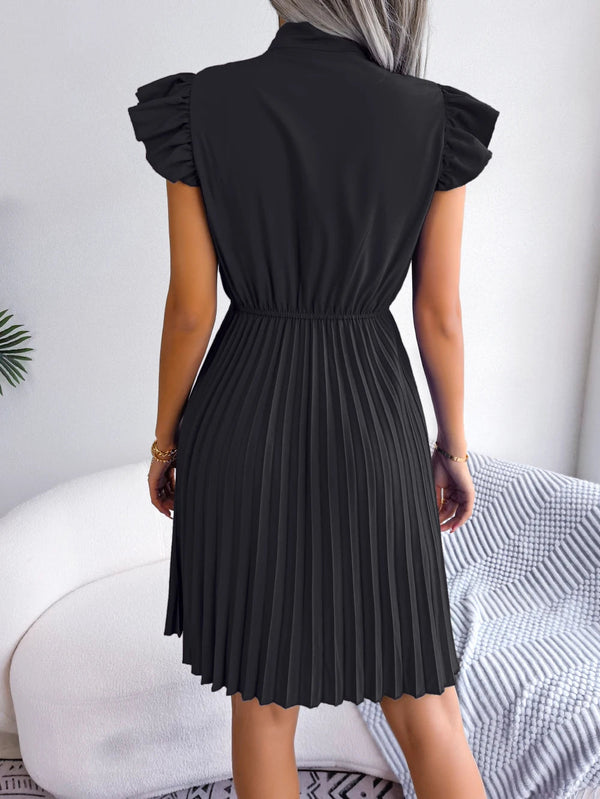 Vrouw in zwarte geplooide jurk met ruches, staand in moderne woonkamer. Elegante damesmode, trendy kleding, stijlvolle outfit voor elke gelegenheid.