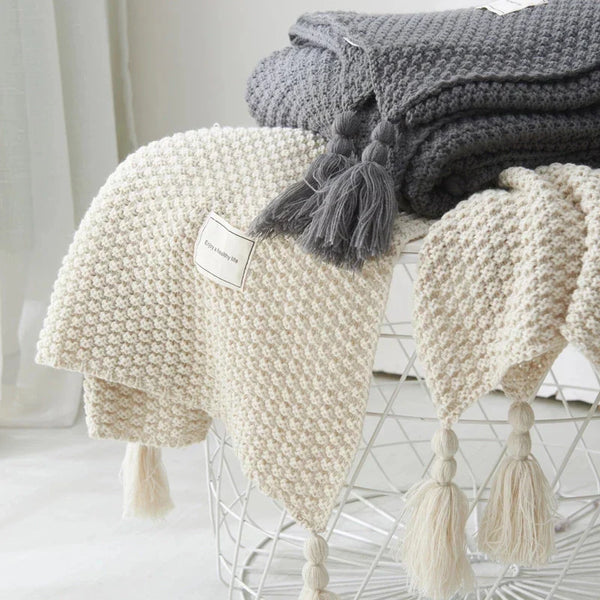 Beige en grijze gebreide dekens met kwastjes op metalen draadmand, stijlvolle woondecoratie, zachte textuur, Scandinavisch interieurontwerp.