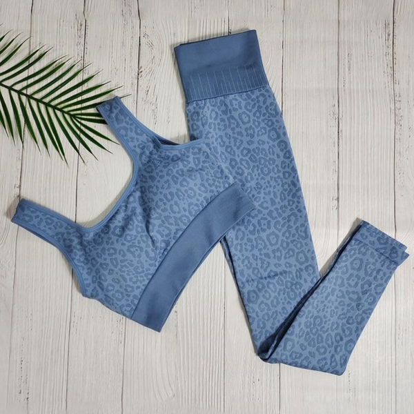 Blauw sportset met luipaardprint, bestaande uit een sportbeha en legging, op witte houten achtergrond met palmblad. Perfect voor yoga en fitness.