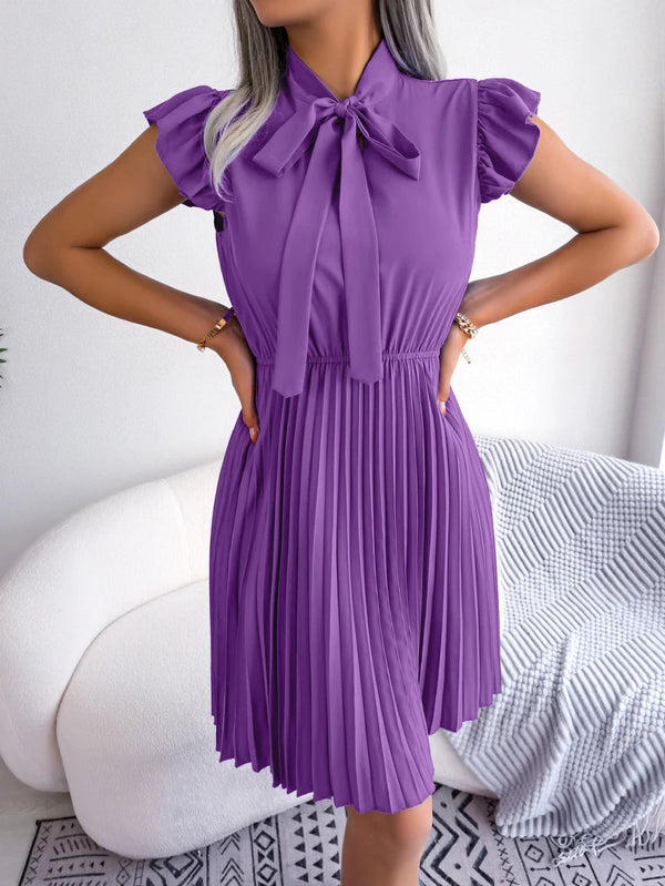 Vrouw in elegante paarse plissé jurk met strik, staand voor witte bank. Modieuze dameskleding, zomerjurk, trendy outfit.