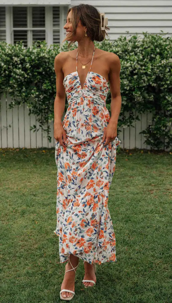 Vrouw in lange, bloemenjurk met halternek, staand op gras voor witte schutting. Zomerse mode, casual elegantie, tuinfeest outfit.
