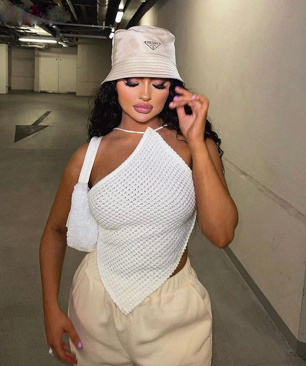 Vrouw in parkeergarage draagt witte asymmetrische top, beige emmerhoed en crèmekleurige broek. Modieuze outfit, stedelijke stijl, zomerlook.