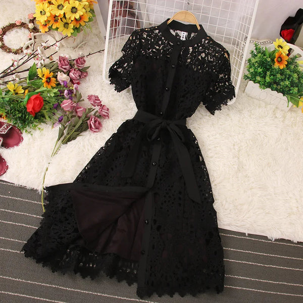 Zwarte kanten jurk met knoopsluiting en strikceintuur, liggend op een witte vloerkleed, omringd door kleurrijke bloemen. Elegante damesmode.