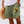 Man in groene korte broek met Kokopelli-design en kleurrijke zoom, houdt een strohoed vast op een strand. Zomerse herenkleding, strandmode.