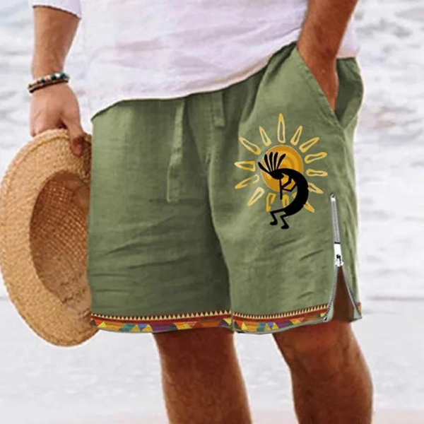 Man in groene korte broek met Kokopelli-design en kleurrijke zoom, houdt een strohoed vast op een strand. Zomerse herenkleding, strandmode.