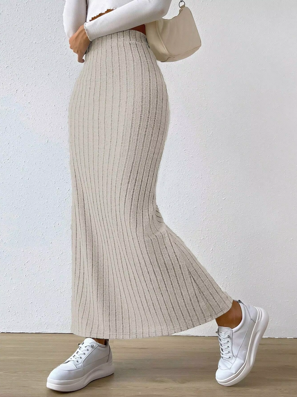 Lange beige geribbelde rok, gecombineerd met witte sneakers en een crèmekleurige handtas, tegen een minimalistische achtergrond. Perfect voor casual mode.
