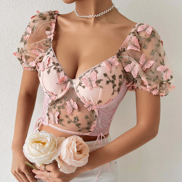 Vrouw draagt roze bloemen crop top met vlindermotief en pofmouwen, gecombineerd met parelketting. Elegante zomermode, trendy dameskleding.