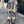 Vrouw in lange, beige en zwarte geometrische printjurk met rieten tas en sandalen, staand op een geplaveide straat voor een smeedijzeren hek.
