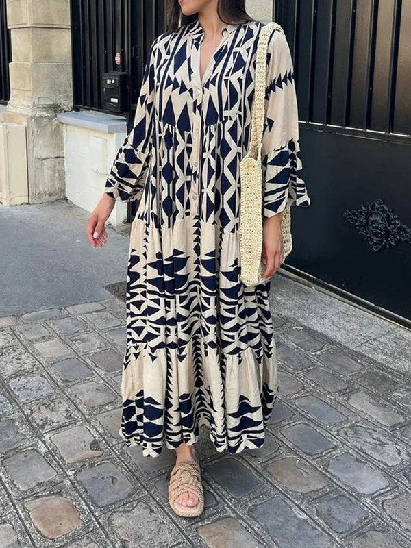 Vrouw in lange, beige en zwarte geometrische printjurk met rieten tas en sandalen, staand op een geplaveide straat voor een smeedijzeren hek.