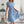 Vrouw in blauwe bloemenjurk met strikceintuur, houdt een rieten tas vast. Stijlvolle zomeroutfit, perfect voor casual en elegante gelegenheden.