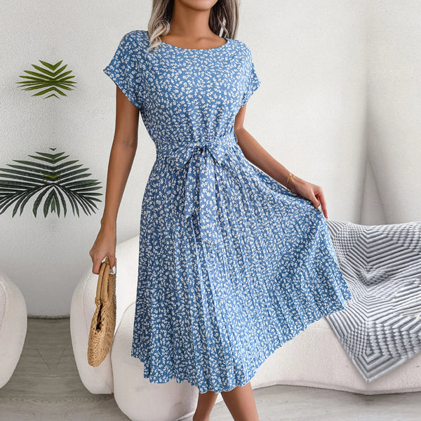 Vrouw in blauwe bloemenjurk met strikceintuur, houdt een rieten tas vast. Stijlvolle zomeroutfit, perfect voor casual en elegante gelegenheden.