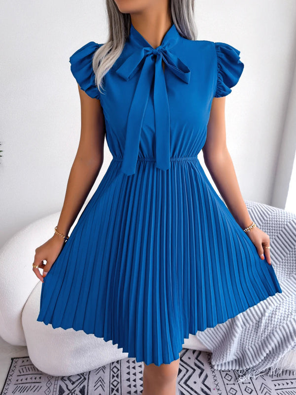 Vrouw in elegante blauwe plisséjurk met strik, korte mouwen, staande in moderne kamer. Mode, dameskleding, stijlvolle jurk, trendy outfit.