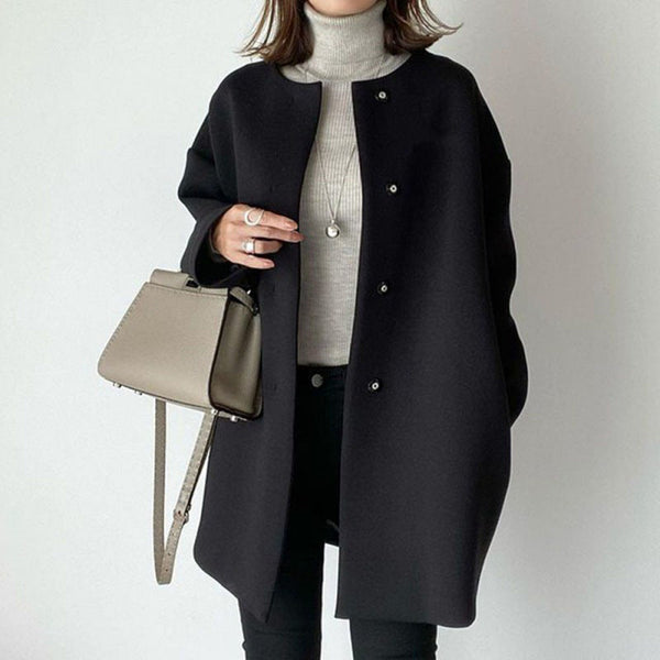 Elegante zwarte damesjas zonder kraag, gecombineerd met een beige handtas en grijze coltrui. Modieuze herfst- en winterkleding.