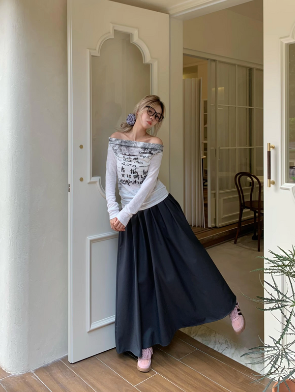 Vrouw in stijlvolle outfit met off-shoulder top en lange zwarte rok, roze sneakers, poseert bij deur. Mode, casual chic, trendy kleding.