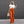 Vrouw in witte gebreide top en oranje flared broek, draagt een beige handtas en zonnebril, poseert voor een grijze achtergrond.