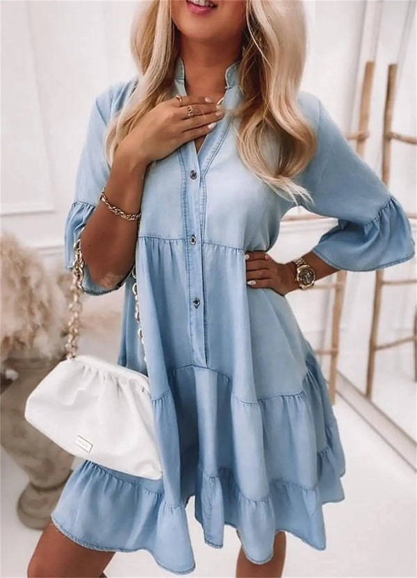 Vrouw in lichtblauwe denim jurk met halflange mouwen en A-lijn patroon, gecombineerd met witte handtas. Trendy damesmode, casual stijl.