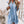 Vrouw in lichtblauwe denim jurk met halflange mouwen, A-lijn patroon, boven de knie, naast decoratieve vaas en ladder. Perfect voor casual lente- of zomeroutfits.