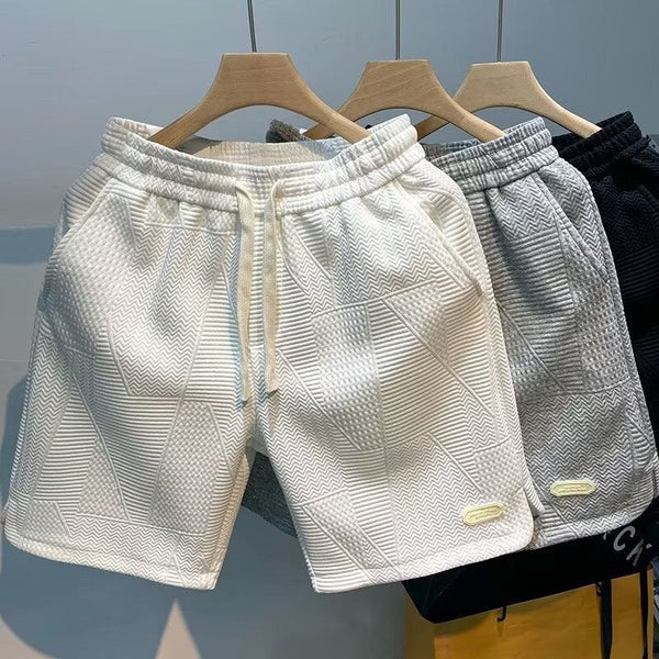 Drie casual herenshorts in wit, grijs en zwart, met elastische tailleband en meerdere zakken, hangend aan houten hangers. Perfect voor zomerse stranddagen.