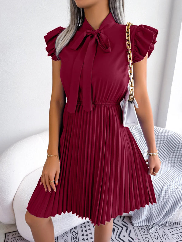 Vrouw in bordeauxrode geplooide jurk met strik en pofmouwen, draagt een witte schoudertas met gouden ketting. Elegante damesmode, trendy outfit.