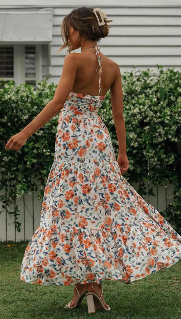 Vrouw in lange, zomerse bloemenjurk met halternek, draaiend in tuin. Elegante mode, casual stijl, perfecte outfit voor warme dagen.