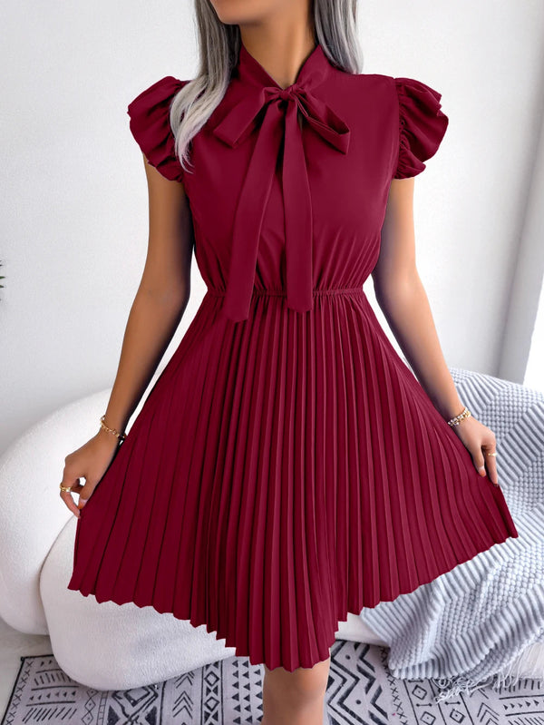 Vrouw in elegante bordeauxrode jurk met plooien en strik, staand in moderne kamer. Perfect voor mode, dameskleding, stijlvolle outfits.