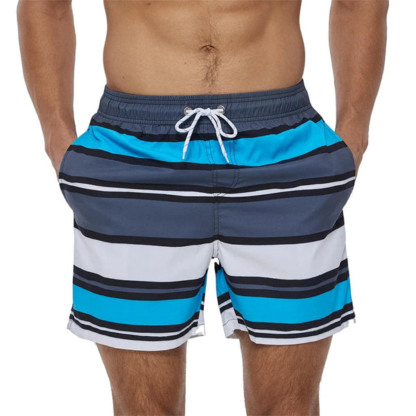 Man in gestreepte zwembroek met blauw, grijs en wit design, handen in zakken. Modieuze heren zwemkleding, strandkleding zomer 2023.