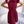 Vrouw in elegante bordeauxrode jurk met plissérok en strikdetail, staand voor een witte bank. Perfect voor mode, dameskleding, stijlvolle outfits.