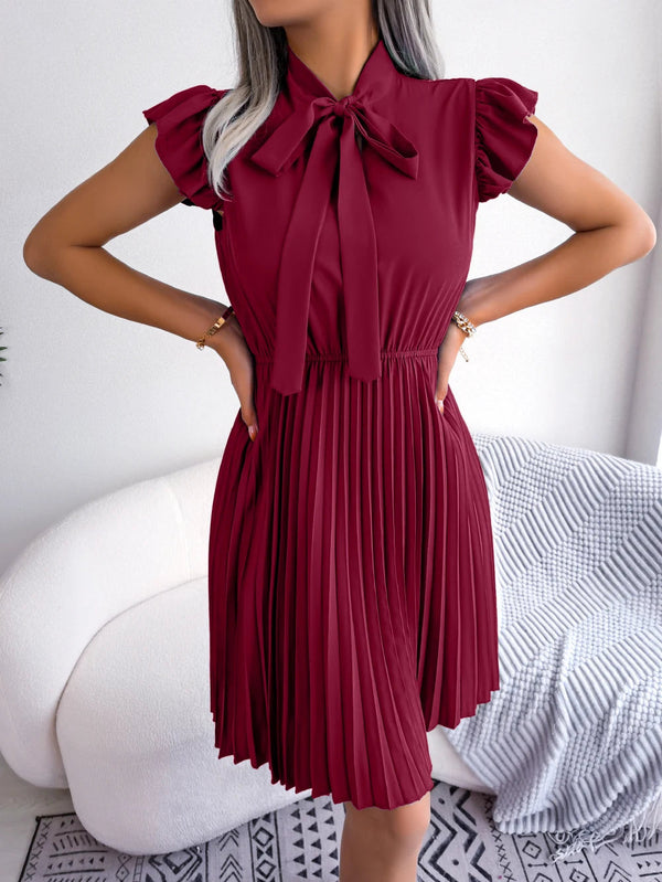 Vrouw in elegante bordeauxrode jurk met plissérok en strikdetail, staand voor een witte bank. Perfect voor mode, dameskleding, stijlvolle outfits.