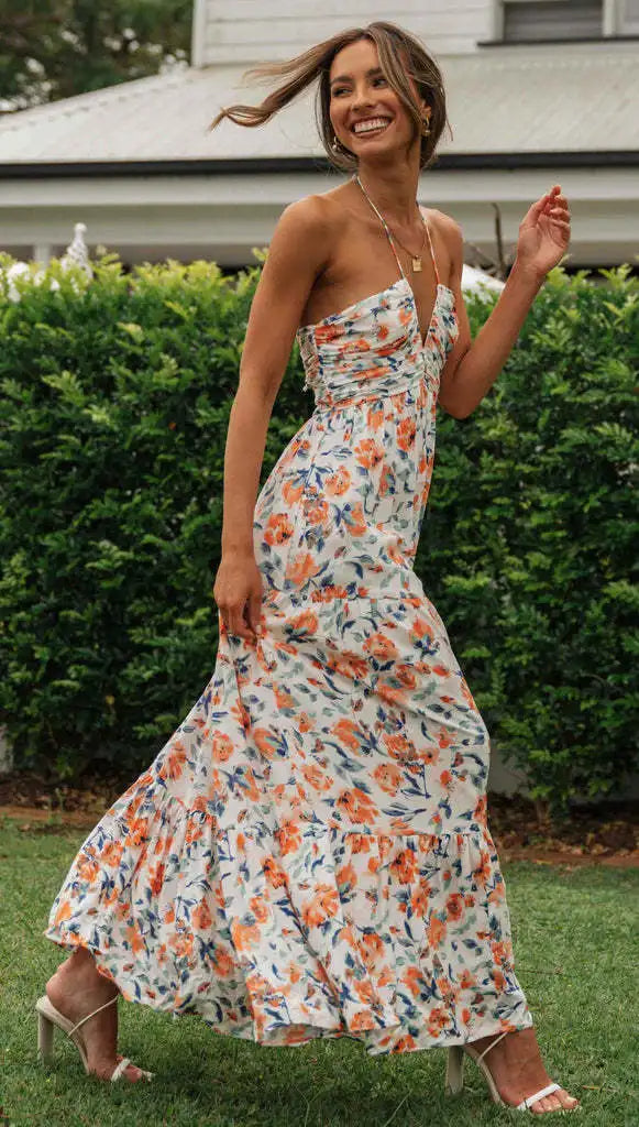 Vrouw in bloemenjurk loopt lachend in tuin, zomermode, elegante maxi-jurk, kleurrijk bloemenpatroon, casual chique stijl, buitenfotografie.