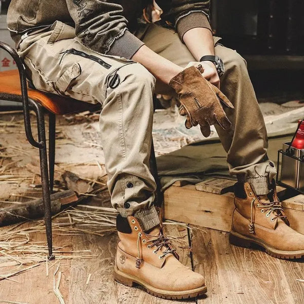 Man in outdoor kleding met beige cargobroek en bruine leren laarzen, zittend op houten stoel. Robuuste handschoenen en industriële omgeving.