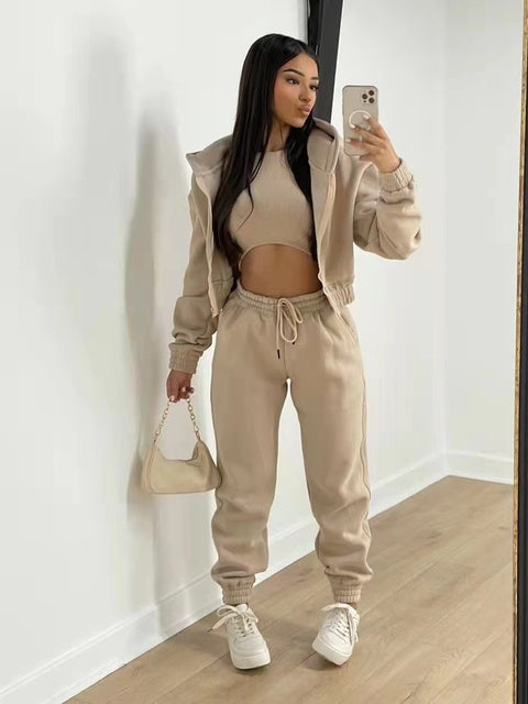 Vrouw in beige joggingpak met crop top, witte sneakers en kleine handtas, poseert voor spiegel. Modieuze casual outfit, trendy dameskleding.