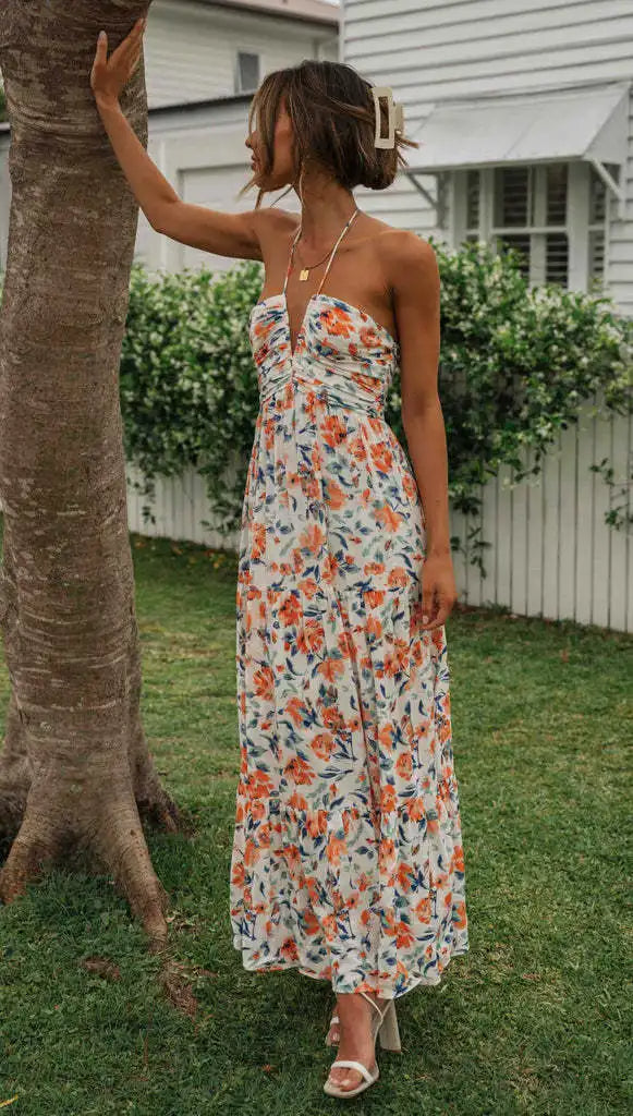 Vrouw in lange, bloemenjurk met halternek, poseert bij boom. Zomerse outfit, casual stijl, tuinomgeving. Perfect voor warme dagen.