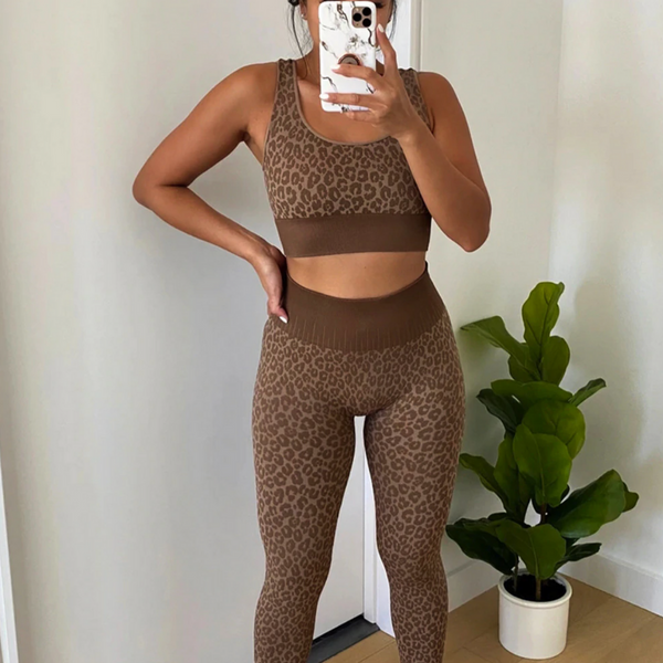 Vrouw in luipaardprint sportset maakt selfie voor spiegel, naast groene kamerplant. Fitnessmode, trendy sportkleding, thuisworkout.