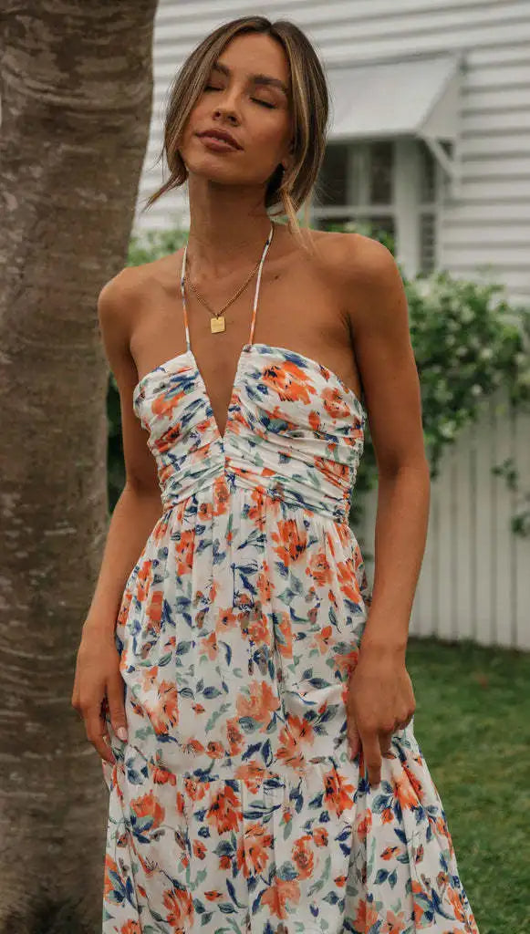 Vrouw in strapless zomerjurk met kleurrijke bloemenprint, staand buiten bij boom en huis. Perfect voor zomermode en casual stijl.