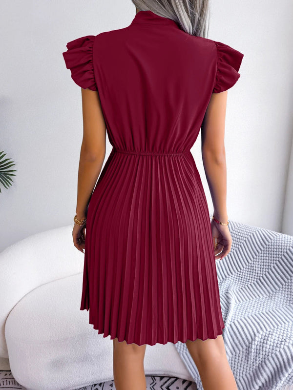 Vrouw in bordeauxrode geplooide jurk met ruches, staand in moderne woonkamer. Elegante damesmode, trendy zomerjurk, stijlvolle kleding.