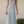 Vrouw in lichtblauwe zomerjurk met halternek, staand in tuin. Elegante maxi-jurk, casual mode, zomerkleding, dameskleding.