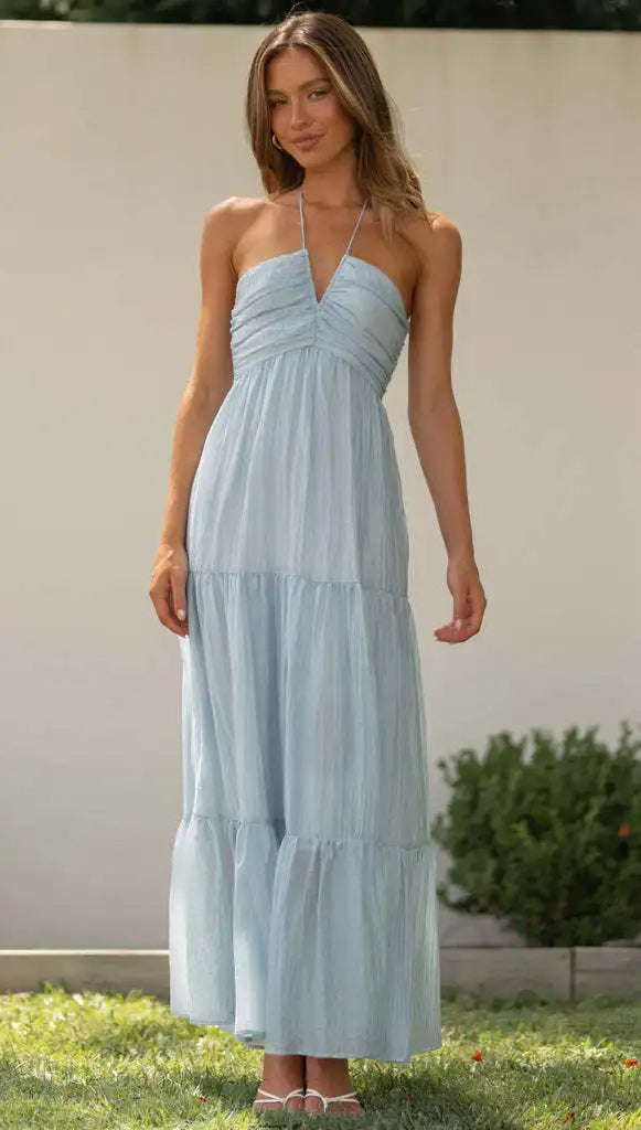 Vrouw in lichtblauwe zomerjurk met halternek, staand in tuin. Elegante maxi-jurk, casual mode, zomerkleding, dameskleding.