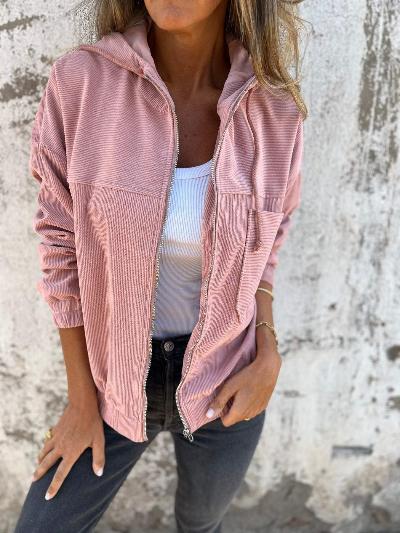 Vrouw draagt roze corduroy jack met capuchon, witte top en zwarte jeans tegen een verweerde muur. Modieuze dameskleding, casual stijl.